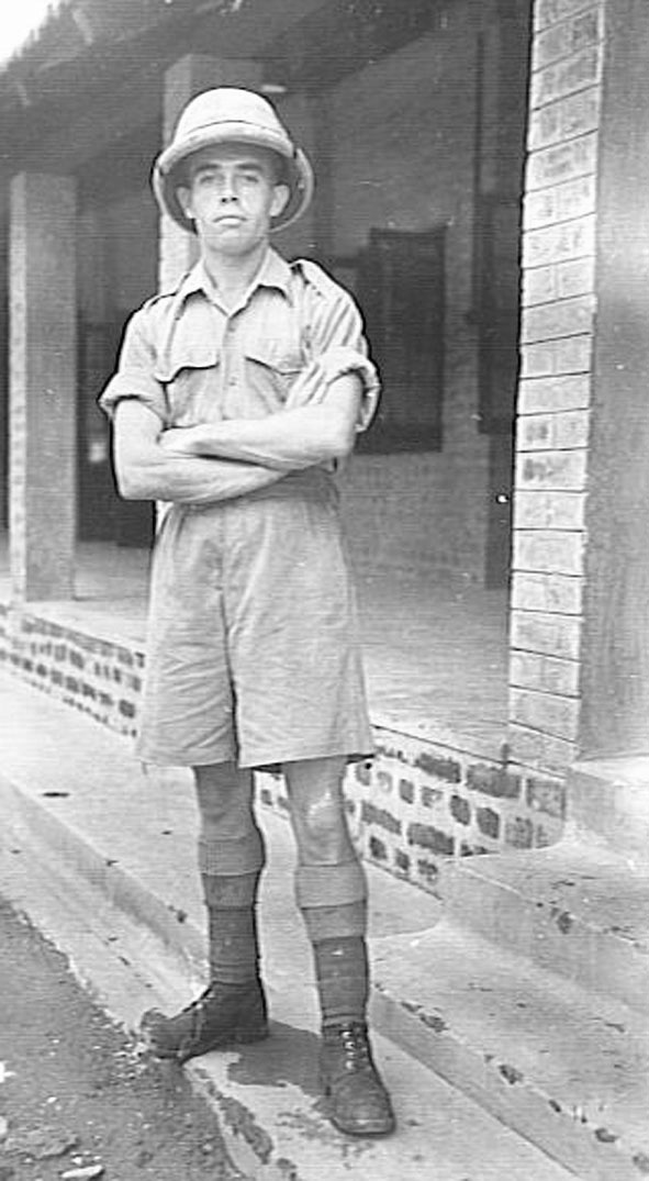 A soldier in tropical kit, pith helmet, shirt and shorts, arms folded, standing on a veranda.