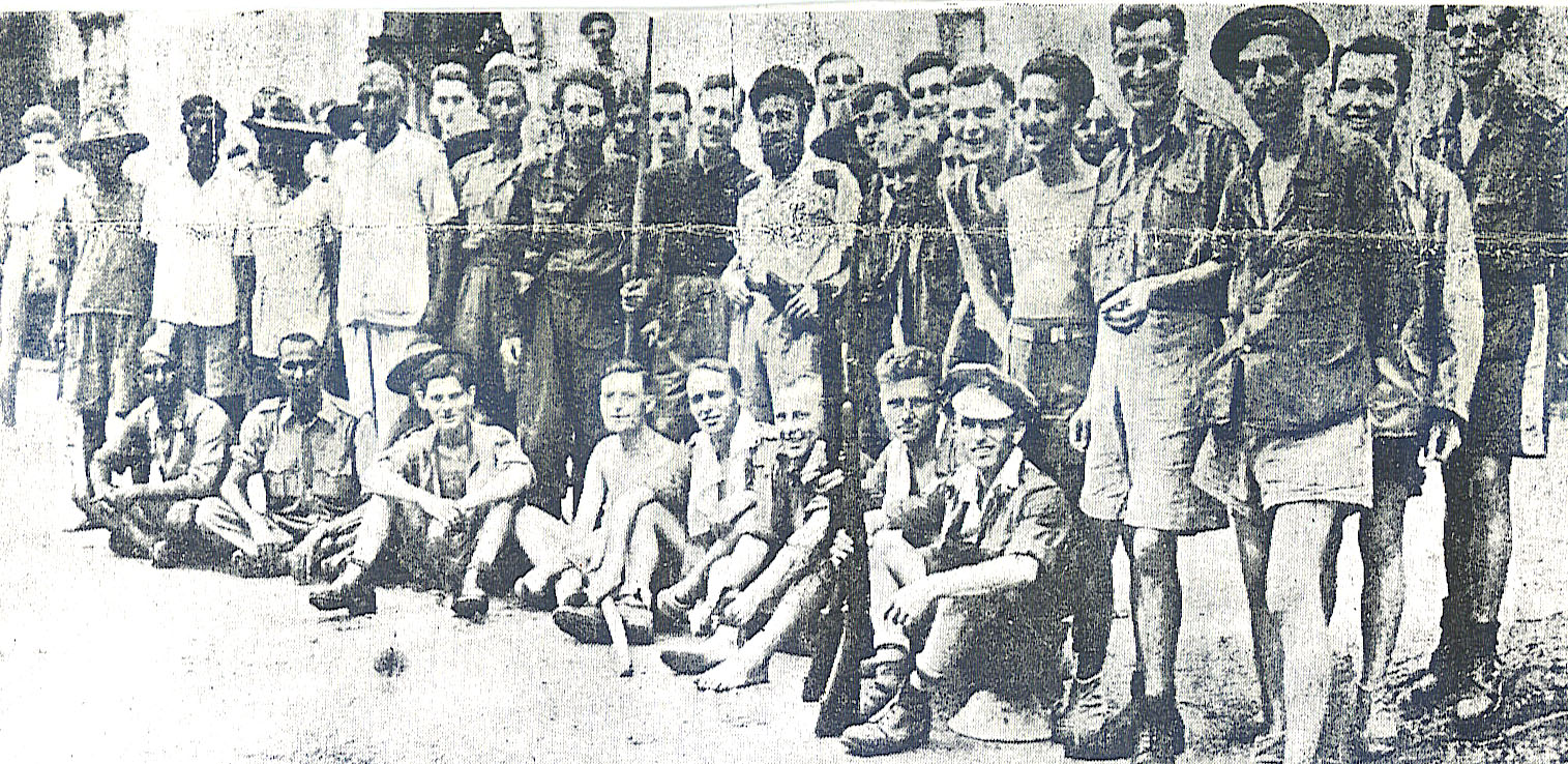 A large group of thin men gathered together outdoors behind a strand of wire after liberation.