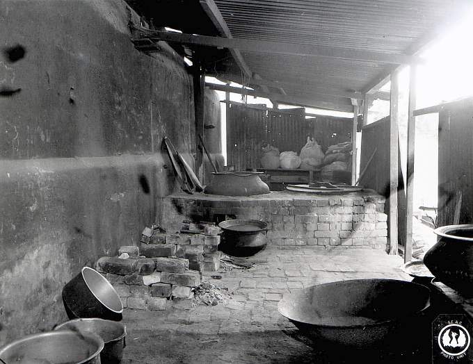 A primitive open cookhouse with large metal pots set over brick fireplaces.