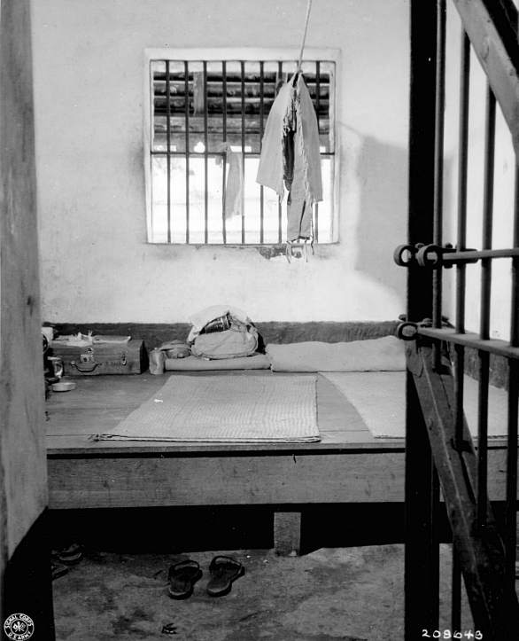 Inside a single cell: a barred window, a sleeping platform with a rolled mat, and sandals on the floor.