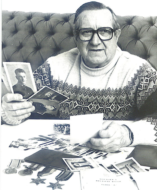 Bill Troughton in later life, seated, holding old photographs, with his medals and papers spread on the table before him.